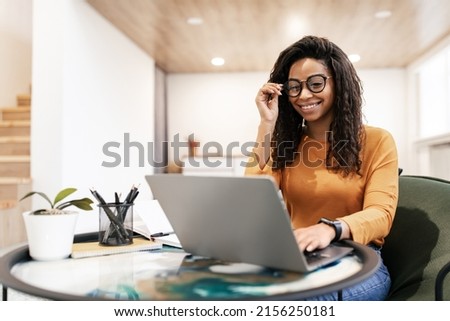 People And Technology. Portrait of young black woman wearing eyeglasses using pc sitting at table on beanbag chair in living room, typing on keyboard. Cheerful lady browsing internet, free copy space Royalty-Free Stock Photo #2156250181