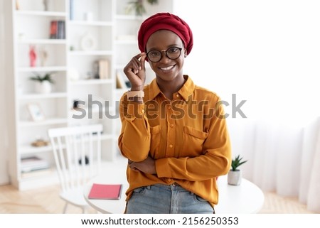 Cute african american young lady in casual outfit and red turban posing at workplace, standing by desk, touching her eyeglasses and smiling at camera, copy space. Career for millennials Royalty-Free Stock Photo #2156250353