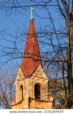 Dark yellow stone church tower with rusty metal roof, leafless spring branches at blue sky background. Church spire with metal cross in Latvia. Lutheran church bell steeple at early spring.