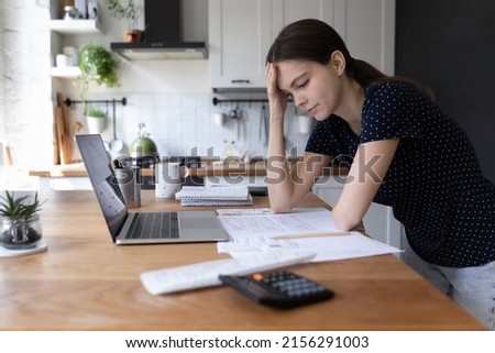 Concerned young woman thinking over domestic paperwork, sitting at laptop computer, papers, calculator at home, touching face, head, analyzing bills, taxes, expenses, financial problems Royalty-Free Stock Photo #2156291003