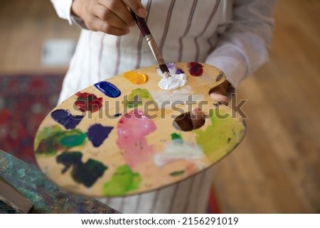 Hands of unknown woman painter holding paintbrush mixing colors on wooden palette, close up cropped view background. Creative hobby, professional occupation, activity for soul, develop talent concept
