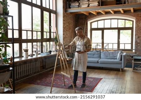 Full-length view in cozy light loft workshop millennial man painter holds palette drawing picture on canvas looks serious and focused engaged in creative activity or hobby for soul, creation concept