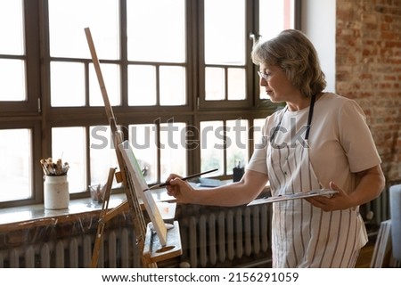 Serious senior female painter stands near easel hold palette and paintbrush creates picture engaged in painting on canvas, creative activity, spend retirement in workshop, enjoy hobby for soul concept