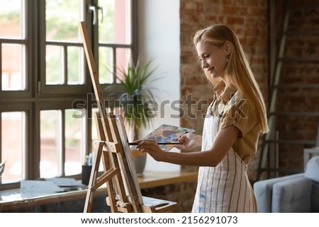 Young beautiful painter woman standing alone in light modern workshop painting picture on canvas with paints looks absorbed, smiles enjoy creative hobby for soul. Education, talent, art-class concept