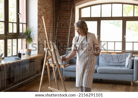 Focused older woman artist standing in loft workshop holds palette and paintbrush, drawing picture on canvas, feeling inspiration enjoy process. Creative hobby on retirement, activity for soul concept