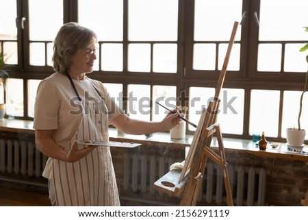 Mature smiling painter woman standing alone in modern workshop painting pictures on canvas with paints looks satisfied, enjoy creative hobby for soul on retirement. Creation, talent, art-class concept