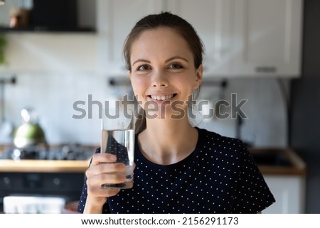 Happy young Caucasian woman drinking fresh pure mineral water in kitchen, holding transparent glass, looking at camera smiling. Home head shot portrait. Healthy lifestyle concept
