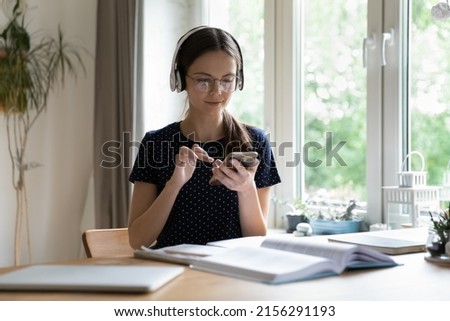 Focused smart student girl in wireless headphones using learning app on mobile phones, watching webinar on smartphone, listening to audio course, studying at open book, copy book at home