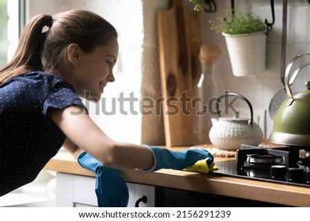 Happy tenant girl using blue protective rubber gloves, rug, cleaning kitchen countertop, cooktop, smiling, enjoying cleanup. Household chores, cleaning service, domestic hygiene concept Royalty-Free Stock Photo #2156291239
