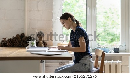 Serious focused smart hardworking student wearing glasses, preparing for exam at home. Young college girl writing essay in copybook, studying book, making notes, summary, doing school homework Royalty-Free Stock Photo #2156291275