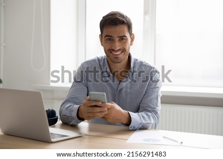 Happy digital gadgets user man using smartphone, laptop at home workplace head shot portrait. Young handsome business man holding mobile phone, sitting at computer, looking at camera Royalty-Free Stock Photo #2156291483