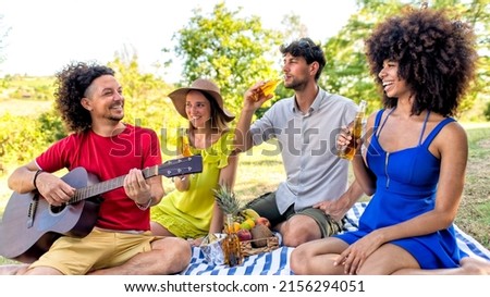 summer holidays outdoor picnic. multiracial group of friends having food and drinking beers laying on a blanket in a park garden. people happy hour enjoying together guitar music. lifestyle concept