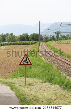 Attention to cyclist road sign post panel at the road side with railway and scenic nature of a countryside. An agriculture fiend and mountain alps in background 
