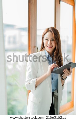 A cheerful, confident, professional, beautiful young Asian businesswoman using a digital tablet stands in an office building representing an online business idea.
