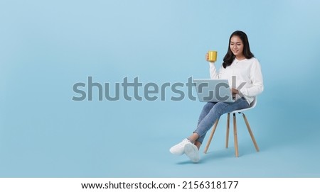 Young woman asian happy smiling in casual white cardigan with denim jeans.While her using computer laptop sitting on white chair and drinking coffee isolate on bright blue background.