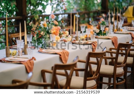 A large, long, decorated, wooden table and chairs, covered with a white tablecloth with dishes, flowers, candles, stands outdoor near the forest in nature. Wedding banquet. lights on background
 Royalty-Free Stock Photo #2156319539