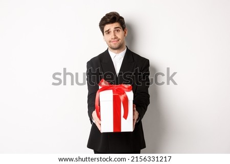Concept of christmas holidays, celebration and lifestyle. Image of handsome boyfriend in black suit, giving you a present, extending hands with gift, standing over white background