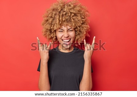 Positive young curly woman shows horn gesture enjoys rock music wears black casual t shirt isolated over vivid red background. Overjoyed curly haired female model demonstrates heavy metal sign