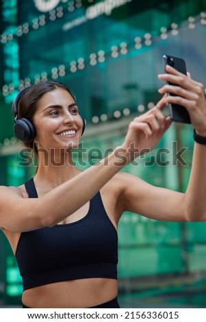 Positive sporty young woman takes selfie via smartphone poses at front camera being in good physical shape dressed in cropped top listens music via headphones poses against blurred background.