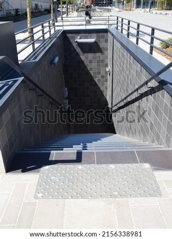 Access staircase to a public car park with tactile strip and visual contrast for the visually impaired in the foreground and the street in the background, perspective view.