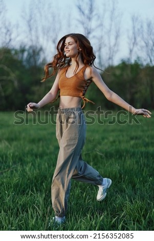 A young woman in a top and green pants and athletic sneakers is jogging on the summer green grass with her hair loose  Royalty-Free Stock Photo #2156352085