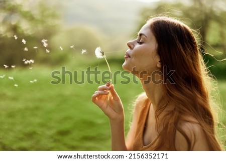 The woman smiles and blows the dandelion seeds into the wind. Summer and sunset light Royalty-Free Stock Photo #2156352171