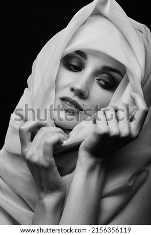 sad beautiful young woman with white headscarf cover her face with luxury makeup on black background looking down, monochrome