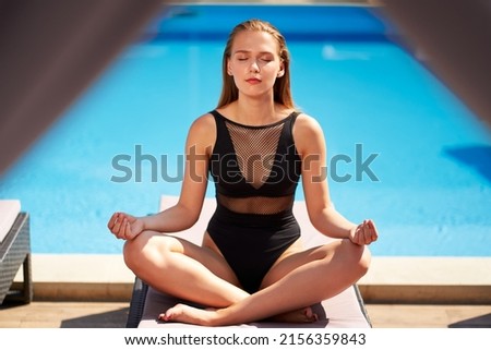 Young athletic woman in bikini meditating by the swimming pool at tropical spa. Fit girl in one piece swimwear practicing zen yoga in resort. Peaceful slim female sitting in lotus pose on beach bed.