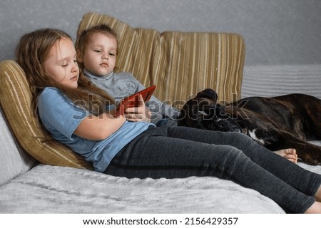two little girls sisters sit on the couch at home and watch the phone, their pets dogs are with them