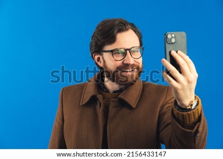 Medium close-up studio shot over blue background. Bearded happy man in his 30s wearing heavy winter brown clothes and glasses holding a smartphone and taking a selfie. High quality photo