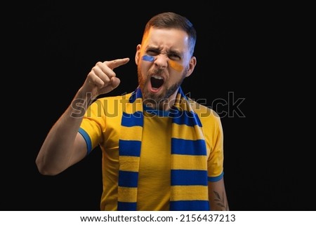 Portrait of excited young soccer supportred man in yellow-blue t-shirt and scarf and painted face, cheering for his favourite team, isolated over black background.