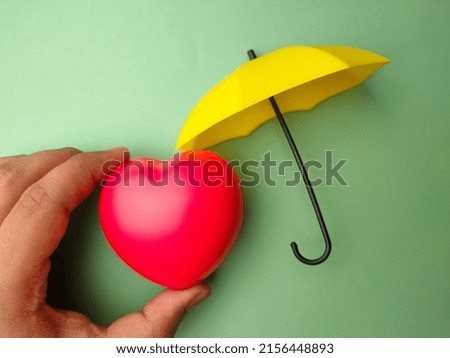 Hand holding red heart with yellow umbrella on a colored background.