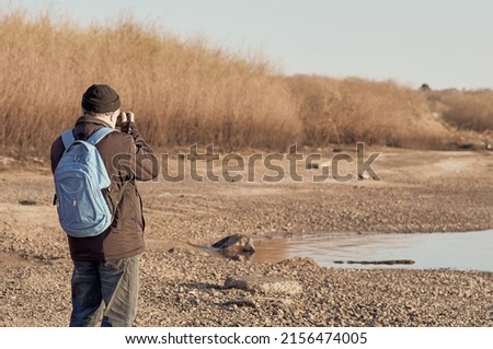 A photographer with a backpack in warm clothes and a hat takes pictures on the shore of a pond. Back view. Spring morning. Selective focus.