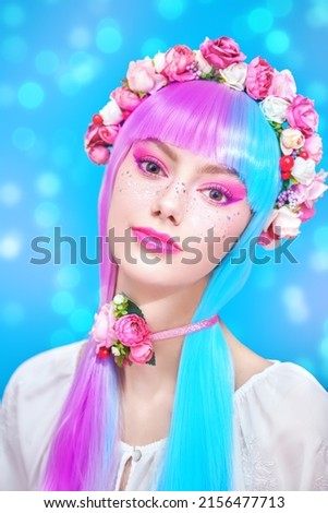 Beauty, makeup and hairstyle. Portrait of a pretty teen girl with bright pink make-up posing in colored violet-blue wig and flower wreath on the head. Blue background with shining lights.