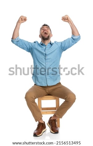 excited man in his forties with arms above head looking up, yelling and celebrating victory while sitting on wooden chair in front of white background  Royalty-Free Stock Photo #2156513495