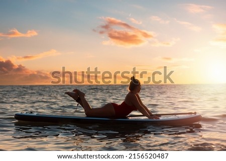 Silhouette of plus sized woman in bikini posing lying on a sup board. Copy space. Sunset sky on the background. The concept of water sports and summer vacation.