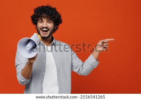 Young bearded Indian man 20s wears blue shirt hold scream in megaphone announces discounts sale Hurry up point on workspace area copy space mock up isolated on plain orange background studio portrait Royalty-Free Stock Photo #2156541265
