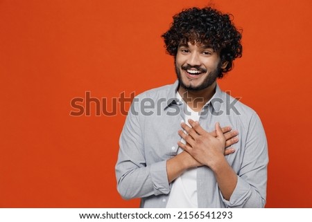 Happy good charming kind fascinating surprised young bearded Indian man 20s years old wears blue shirt ask who me oh it so sweet put hands on chest isolated on plain orange background studio portrait Royalty-Free Stock Photo #2156541293