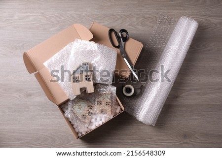 Open box with decorative house figures, adhesive tape, scissors, foam peanuts and bubble wrap on wooden table, flat lay Royalty-Free Stock Photo #2156548309