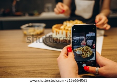 Young woman taking pictures of a cake in the kitchen. Female baker capturing photos of pastry items with her mobile phone.