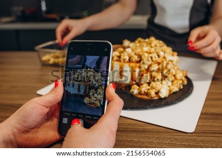 Young woman taking pictures of a cake in the kitchen. Female baker capturing photos of pastry items with her mobile phone.