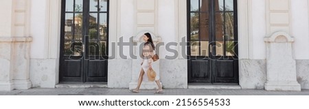 side view of trendy woman walking along white building with black doors, banner