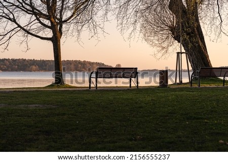 Empty park bench in front of an idyllic lake. The sun is going down and the sky is glowing in orange color. Tranquil scenery without people. Landscape with a resting place in a lakeside area. Royalty-Free Stock Photo #2156555237