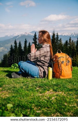 A young, slender girl in a plaid shirt and jeans with an orange backpack meditates sitting on the green grass against the backdrop of mountains.