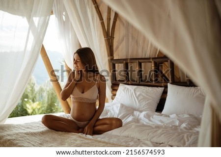 Attractive pregnant woman is sitting in bed and holding her belly. The girl in the bamboo house. Last months of pregnancy.