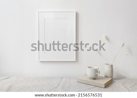 Neutral breakfast still life scene. White wooden picture frame mockup. Vase with dry lagurus bunny tail grass. Cup of coffee on book. Linen table cloth, white wall. Scandinavian interior. Boho style.