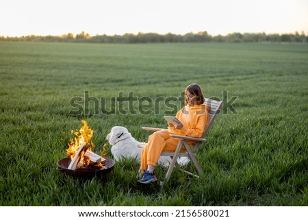Young person sitting on chair near the fireplace and using digital device while relaxing with pet on green field during the sunset. Concept of recreation on nature and freelance Royalty-Free Stock Photo #2156580021
