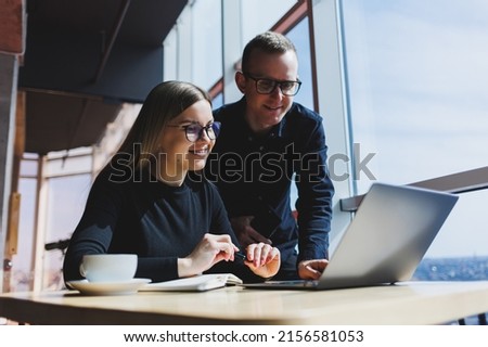 Smiling young male and female manager sitting at table, looking at computer screen, explaining enterprise software or working on online project with colleagues.