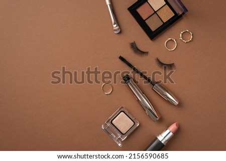 Make-up concept. Top view photo of eyeshadow palette makeup brush gold rings lipstick mascara and false eyelashes on isolated brown background with copyspace