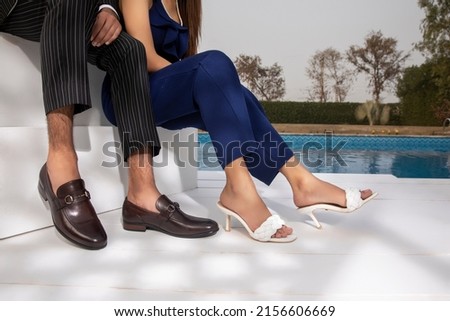 The girl and the boy are sitting on a bench near the pool, the girl is wearing a blue dress, white high heels, and the boy is wearing a black pants shirt and black shoe, there is a pool. Royalty-Free Stock Photo #2156606669